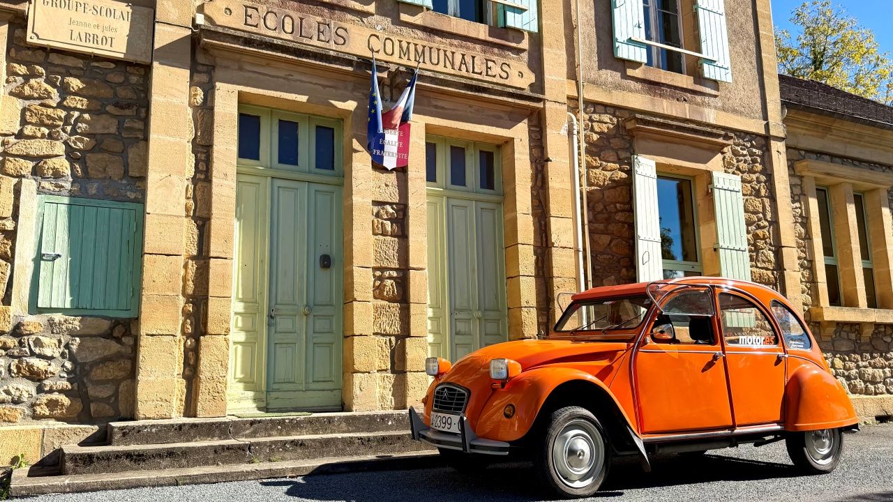 Un Citroën 2CV naranja junto a un antiguo colegio francés.