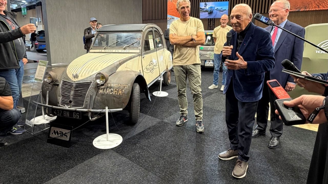 Varias personas escuchan hablar al anciano publicista Jacques Séguéla junto a un Citroën 2CV gris y blanco.