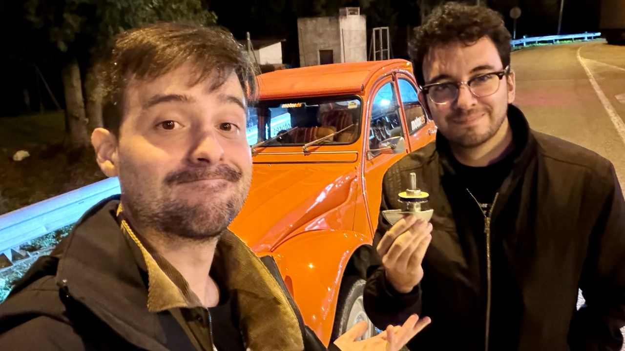 Dos hombres junto a un Citroën 2CV naranja; el de la derecha sostiene el tirador de la puerta, que se ha caído.