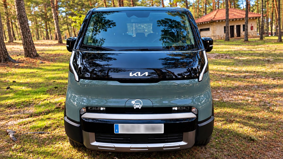 Frontal de una furgoneta Kia de color verde y negro estacionada sobre un terreno con hierba en un entorno boscoso a la luz del día.